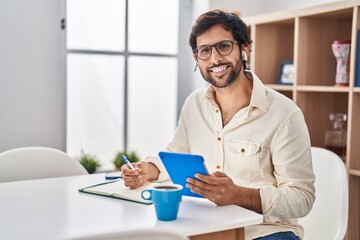 Young hispanic man writing on notebook using touchpad at home