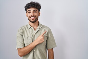 Arab man with beard standing over white background cheerful with a smile on face pointing with hand and finger up to the side with happy and natural expression
