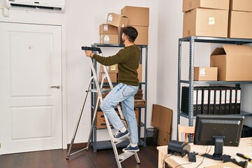 Young arab man ecommerce business worker holding package standing on ladder at office