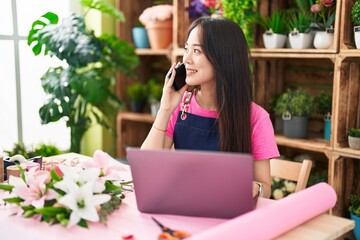 Young chinese woman florist talking on smartphone using laptop at flower shop - Powered by Adobe