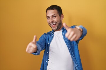 Hispanic man standing over yellow background approving doing positive gesture with hand, thumbs up smiling and happy for success. winner gesture.