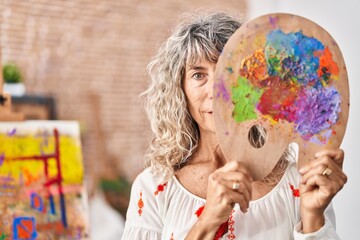 Fototapeta premium Middle age woman holding painter palette covering face relaxed with serious expression on face. simple and natural looking at the camera.