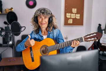 Middle age woman playing classic guitar at music studio smiling happy and positive, thumb up doing excellent and approval sign