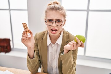 Young caucasian woman working at the office eating snack angry and mad screaming frustrated and...