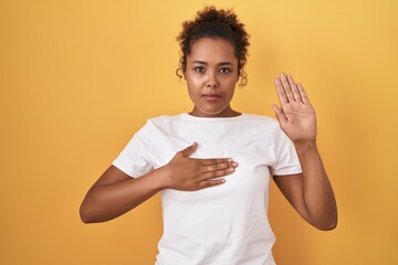 Young hispanic woman with curly hair standing over yellow background swearing with hand on chest...