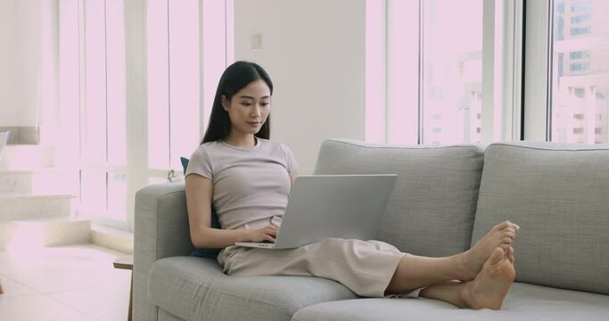 Attractive focused Asian woman spend free time on internet seated on cozy sofa with laptop, do telework or e-shopping, using wireless device for remote work, freelance job at home. Modern tech usage
