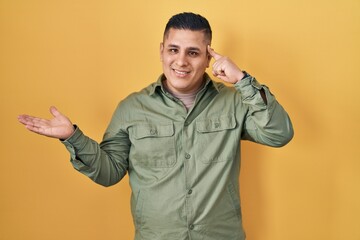 Hispanic young man standing over yellow background confused and annoyed with open palm showing copy...