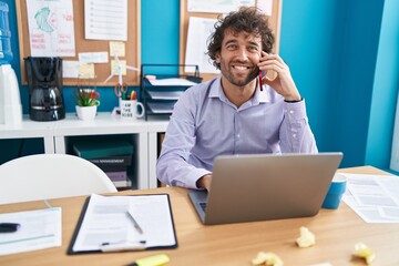 Young hispanic man business worker using laptop talking on smartphone at office