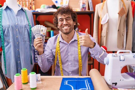 Hispanic young man dressmaker designer holding dollars smiling happy and positive, thumb up doing excellent and approval sign
