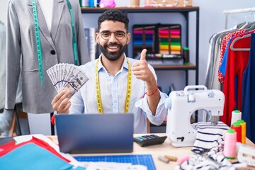 Hispanic man with beard dressmaker designer holding dollars smiling happy and positive, thumb up doing excellent and approval sign