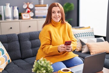 Young beautiful plus size woman using smartphone and laptop sitting on sofa at home