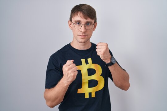 Caucasian blond man wearing bitcoin t shirt ready to fight with fist defense gesture, angry and upset face, afraid of problem