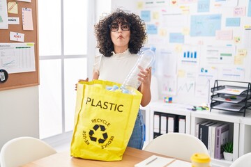 Young middle east woman holding recycling bag with plastic bottles at the office making fish face...