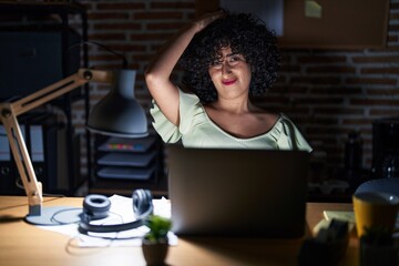 Young brunette woman with curly hair working at the office at night confuse and wondering about...