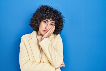 Young brunette woman with curly hair standing over blue background thinking looking tired and bored with depression problems with crossed arms.