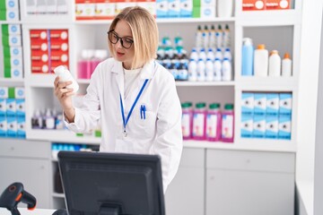 Young blonde woman pharmacist using computer holding pills bottle at pharmacy