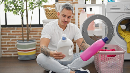 Young hispanic man washing clothes choosing detergent bottle at laundry room