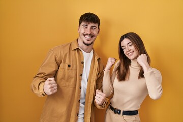 Young hispanic couple standing over yellow background very happy and excited doing winner gesture...