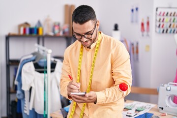 Young hispanic man tailor smiling confident writing on notebook at tailor shop