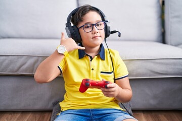 Young hispanic kid playing video game holding controller wearing headphones smiling doing phone gesture with hand and fingers like talking on the telephone. communicating concepts.