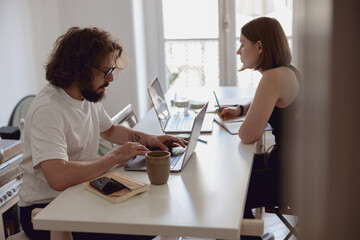 Married couple freelancers sit at table in living room with laptops and working from home office