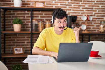 Hispanic man wearing call center agent headset working from home pointing thumb up to the side smiling happy with open mouth