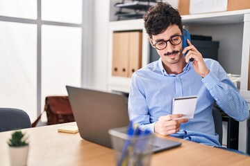 Young caucasian man business worker talking on smartphone reading notebook at office