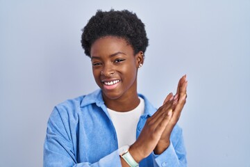 African american woman standing over blue background clapping and applauding happy and joyful,...