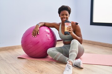 African american woman drinking smoothie sitting on yoga mat at sport center