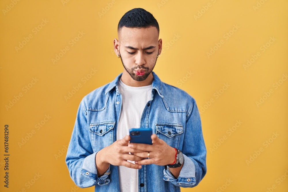 Sticker Young hispanic man using smartphone typing message puffing cheeks with funny face. mouth inflated with air, catching air.