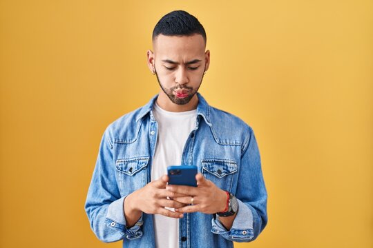 Young hispanic man using smartphone typing message puffing cheeks with funny face. mouth inflated with air, catching air.