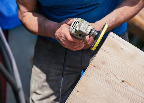 Man polishing wooden chest with old angle grinder during sunny day, closeup detail to hands without gloves