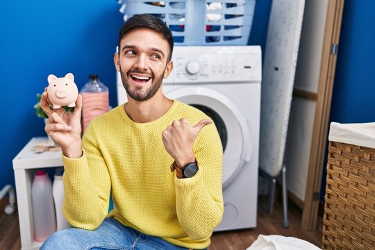 Hispanic man doing laundry holding piggy bank pointing thumb up to the side smiling happy with open mouth