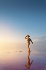 Fototapeta na wymiar In the evening, on the lake, a girl walks on the water, with a reflection. A young woman stands barefoot in a pastel pink powdery lake with white crystallized salt