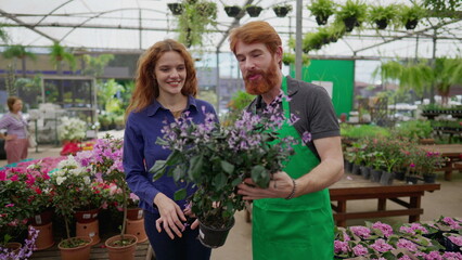 Man helping customer to select plant at Local Business Flower Shop. A redhead male person with green apron assisting a female customer