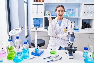 Young brunette woman working at scientist laboratory smiling with happy face winking at the camera doing victory sign. number two.