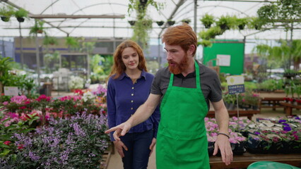 Fototapeta na wymiar Redhead Man in Green Apron Assisting Female Customer in Local Flower Shop
