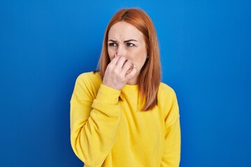 Young woman standing over blue background smelling something stinky and disgusting, intolerable...