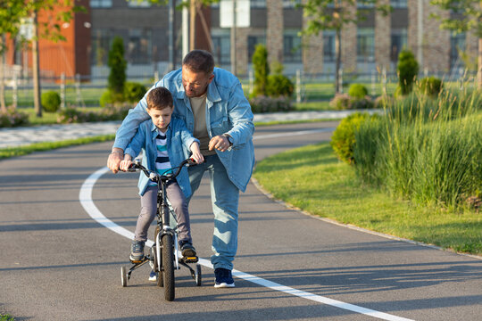 Father Teaching Son Riding Bike. Father Helping Excited Son To Ride A Bicycle In American Neighborhood. Child Learning To Ride Cycle With His Dad. Fathers Day.