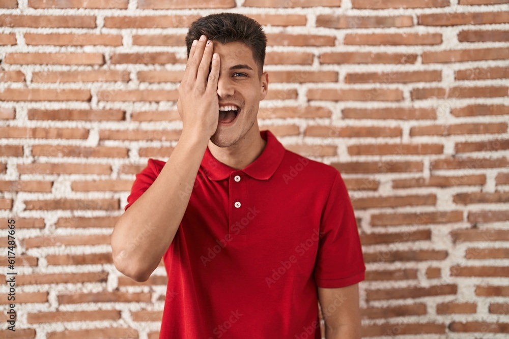 Sticker young hispanic man standing over bricks wall covering one eye with hand, confident smile on face and