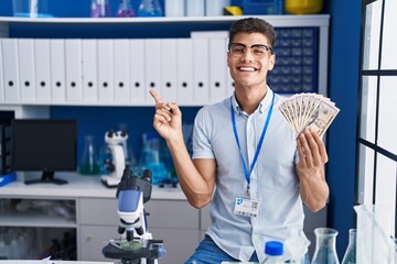 Young hispanic man working at scientist laboratory holding dollars smiling happy pointing with hand...