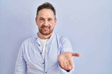 Middle age caucasian man standing over blue background smiling cheerful offering palm hand giving assistance and acceptance.