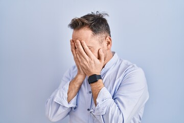 Middle age caucasian man standing over blue background with sad expression covering face with hands...