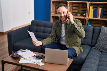 Young caucasian man talking on smartphone reading document at home