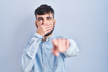 Young hispanic man with beard standing over blue background laughing at you, pointing finger to the camera with hand over mouth, shame expression
