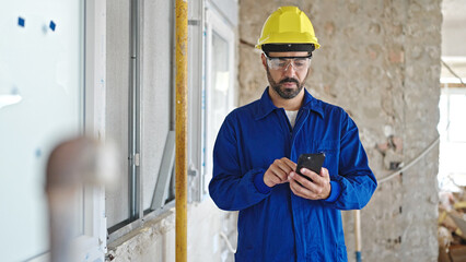 Young hispanic man worker wearing hardhat using smartphone at construction site