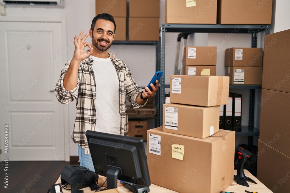 Sticker Young hispanic man with beard working at small business ecommerce with smartphone doing ok sign with fingers, smiling friendly gesturing excellent symbol