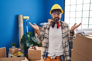 Young hispanic man with beard working at home renovation clueless and confused expression with arms...
