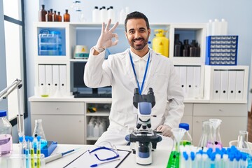 Fototapeta premium Young hispanic man with beard working at scientist laboratory smiling positive doing ok sign with hand and fingers. successful expression.
