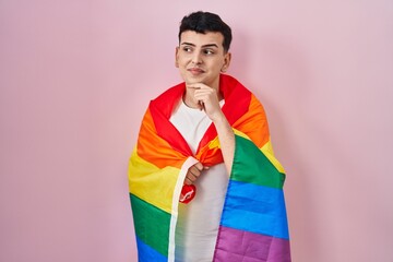 Non binary person holding rainbow lgbtq flag serious face thinking about question with hand on chin, thoughtful about confusing idea